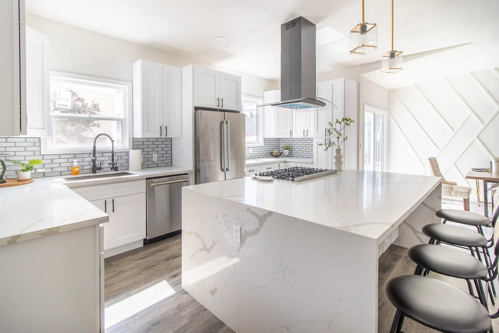 Bright, modern kitchen featuring a large white marble island with a gas cooktop, sleek white cabinets, stainless steel appliances, and bar stools, in a sunlit dining area.