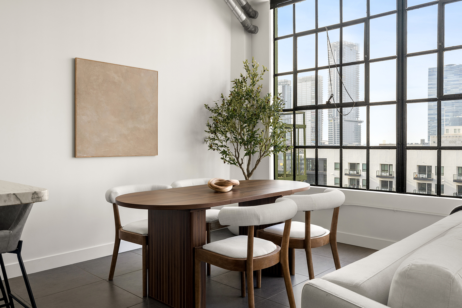 Bright dining area with a modern wooden table surrounded by upholstered chairs, a potted olive tree, and large industrial-style windows revealing a city skyline.