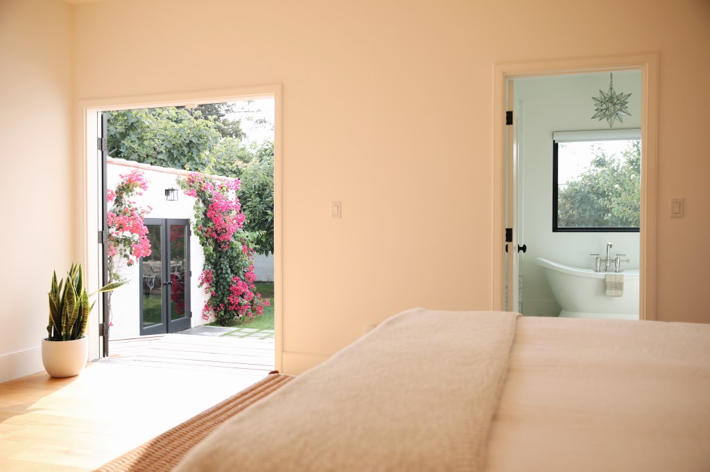 Cozy bedroom with a soft white bedspread, an open door leading to a patio adorned with pink bougainvillea, and a bathroom entrance showcasing a bathtub and outdoor trees visible through the window.