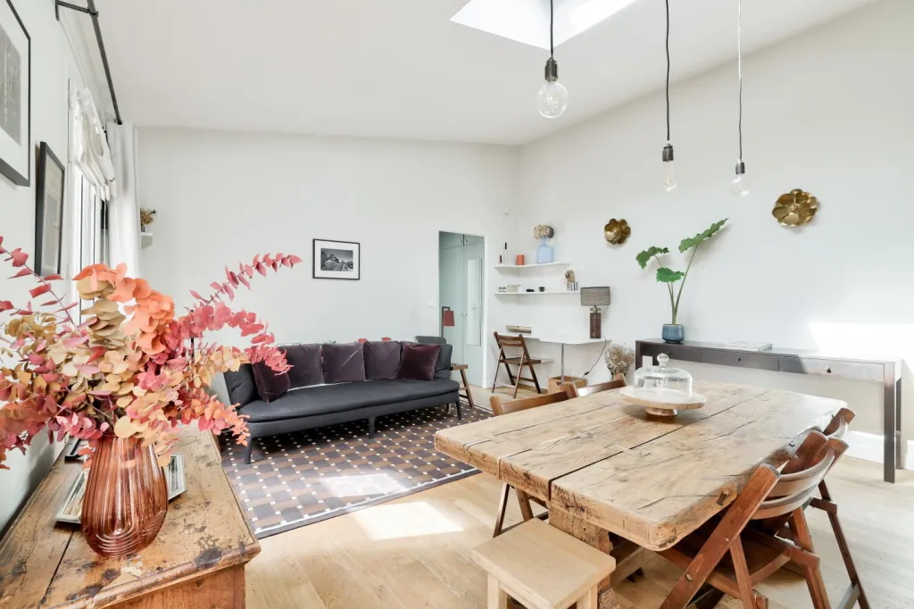 A bright and airy living-dining space features rustic wooden furniture, modern lighting, and colorful dried flowers for a cozy yet stylish vibe.