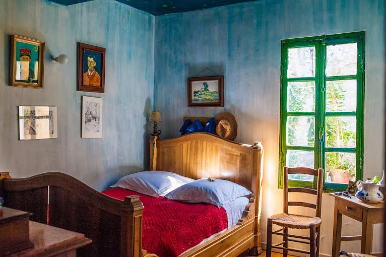 Cozy bedroom with a wooden bed frame, red blanket, and blue pillows, surrounded by artwork on blue walls and sunlight streaming through a green-framed window.