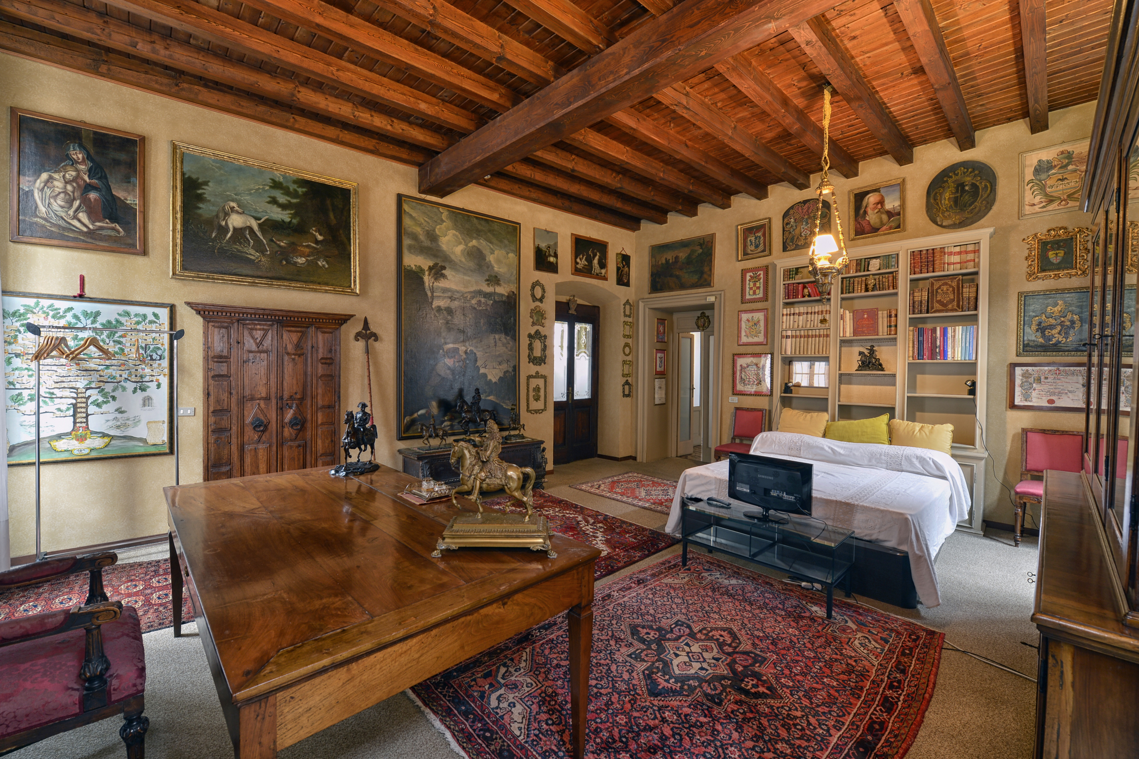 A beautifully decorated room featuring a wooden beamed ceiling, antique furniture, and walls adorned with colorful artwork and framed paintings. A bed with white sheets is situated near a bookshelf filled with books and collectibles.