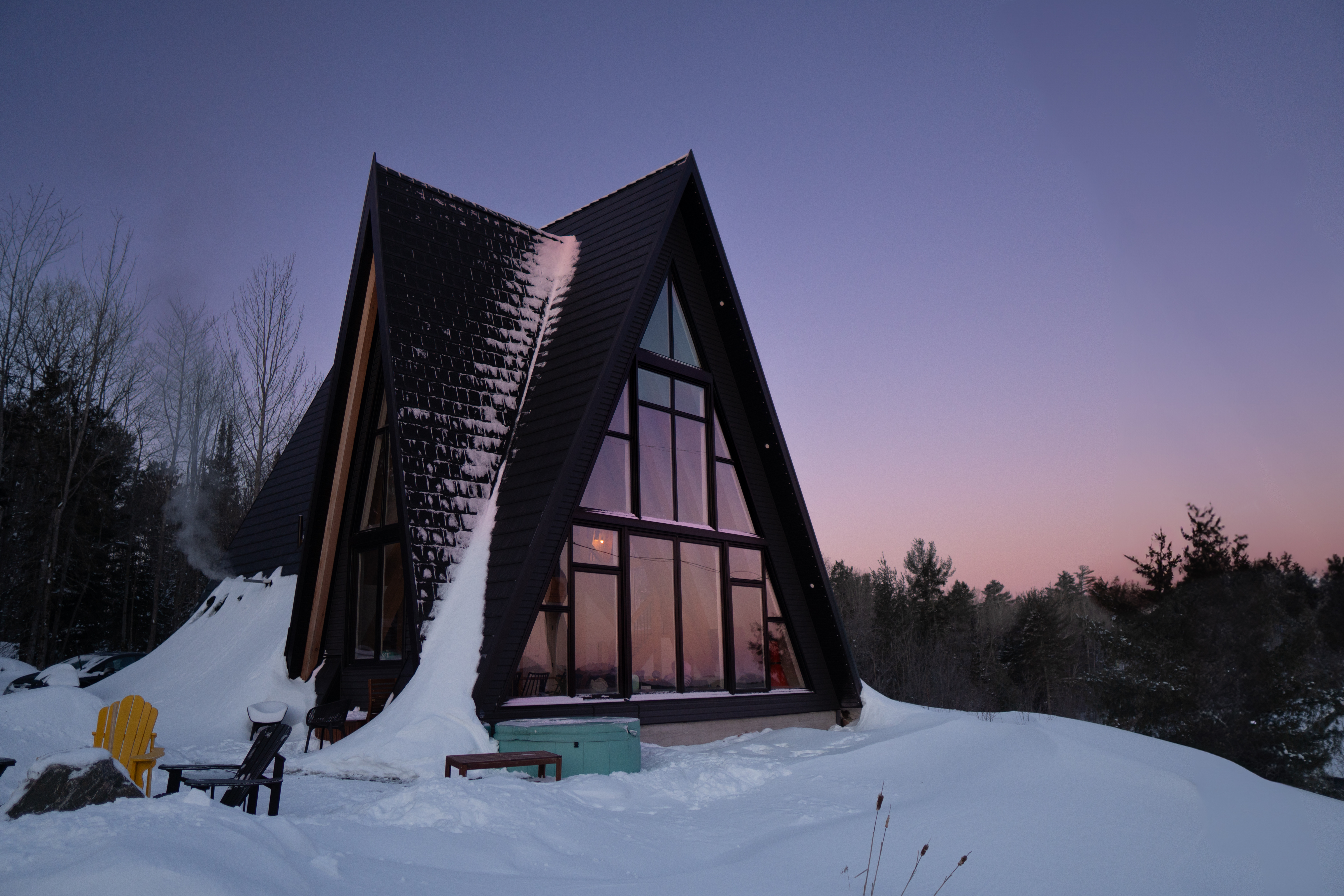 Black A-frame cabin covered in snow with a pink sunset in the background.