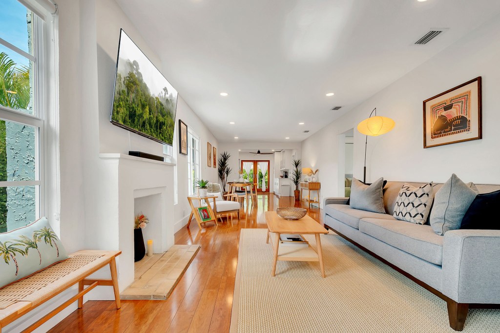 A bright and spacious living room with a modern gray sofa, wooden coffee table on a beige rug, a wall-mounted TV above a white fireplace, and large windows letting in natural light with views of greenery outside.