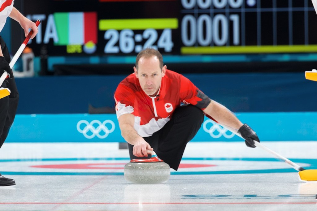 Athlete in a red and white uniform sliding a curling stone on ice during a match, with Olympic rings visible in the background.