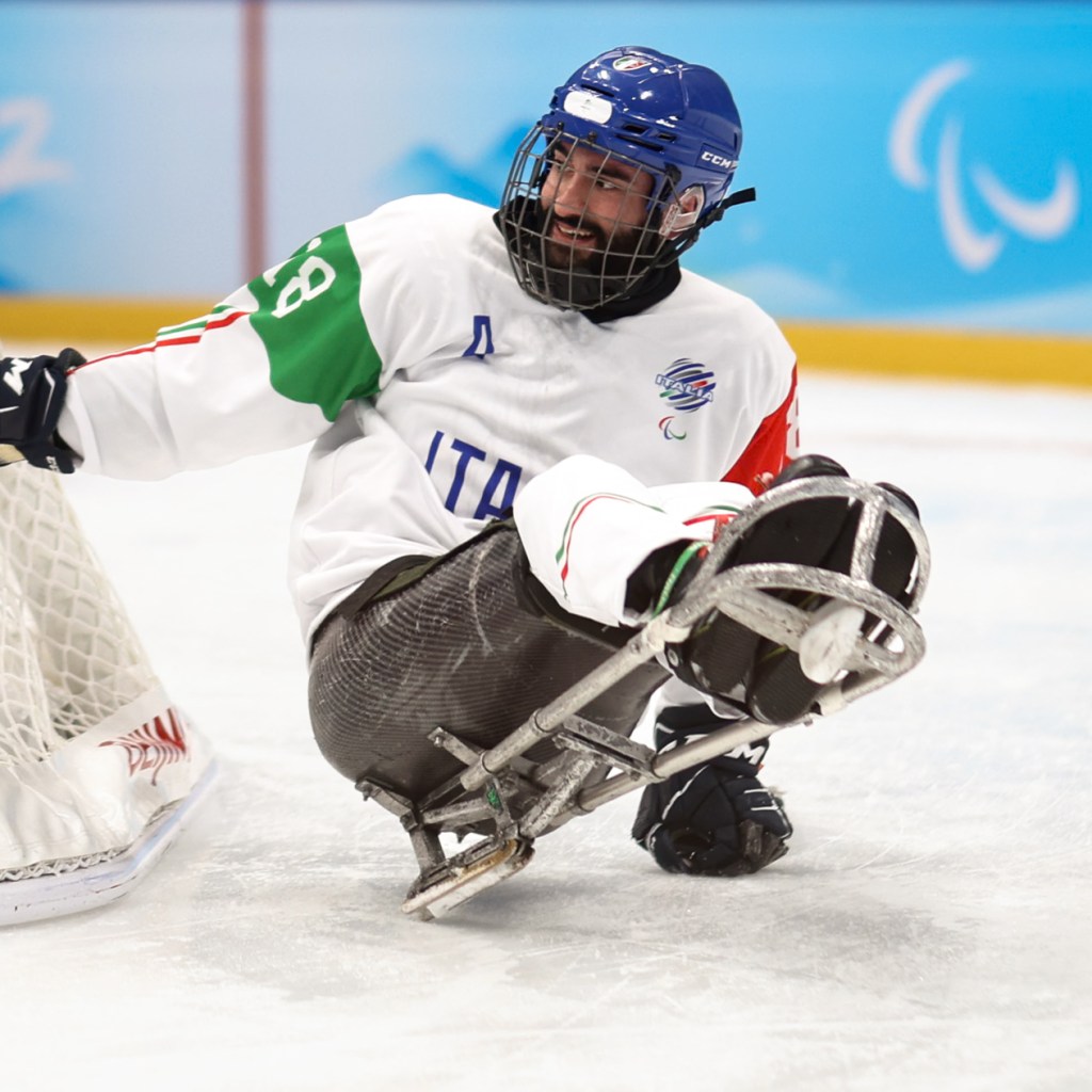 An Italian para ice hockey player wearing a white jersey with 'ITA' lettering and a blue helmet in action on an ice rink, using a sled.