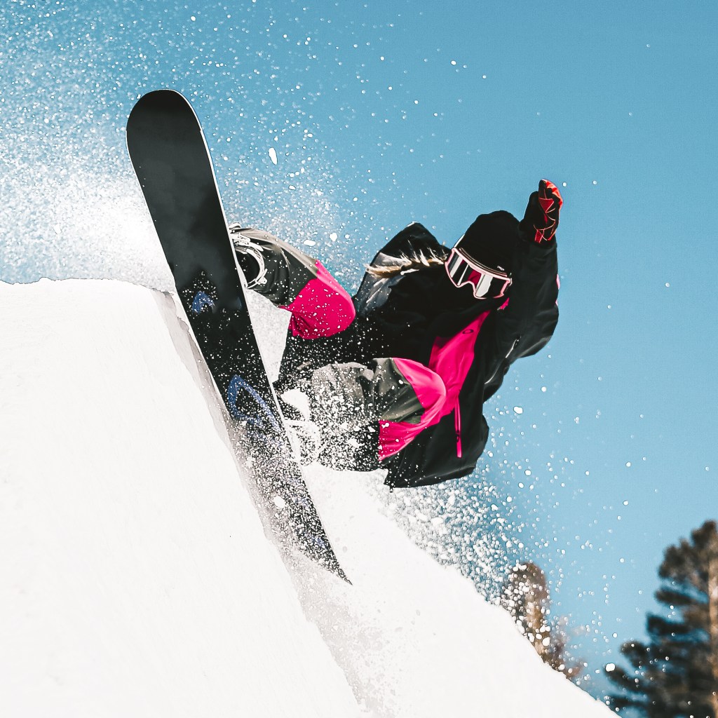 A snowboarder in black and pink gear jumping off a snow-covered slope, with snow spraying into the air against a bright blue sky.