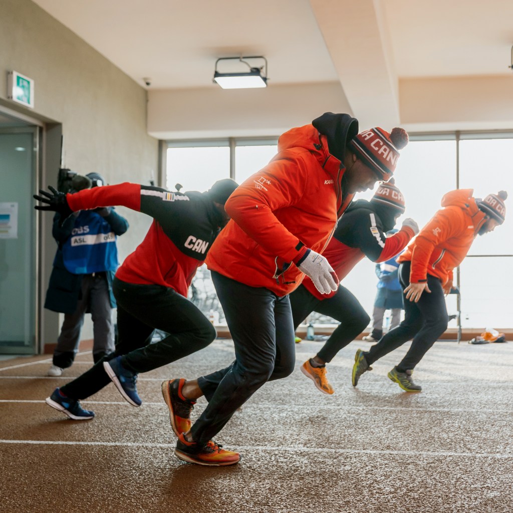 Four team members in athletic gear crouched in a starting position on an indoor track, preparing for practice.