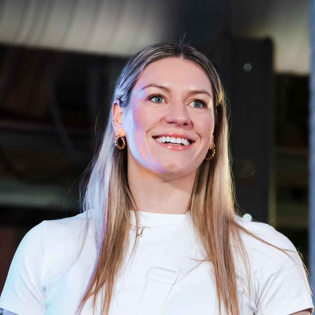 Individual with straight blonde hair and small gold accessories, dressed in a white outfit with minimalistic lighting and industrial background.