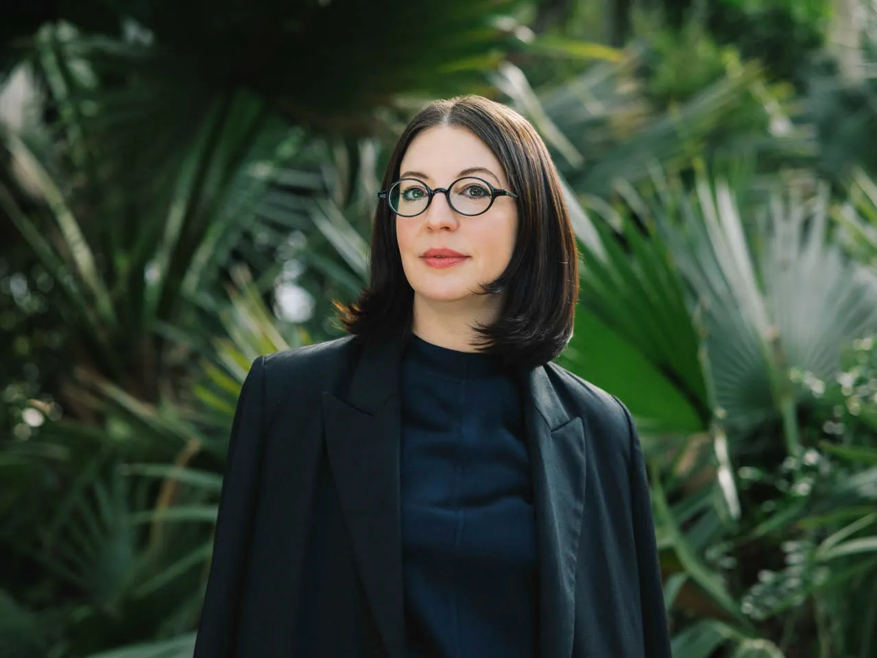 A person wearing a black blazer and dark shirt, standing in front of lush green palm leaves in a garden setting.