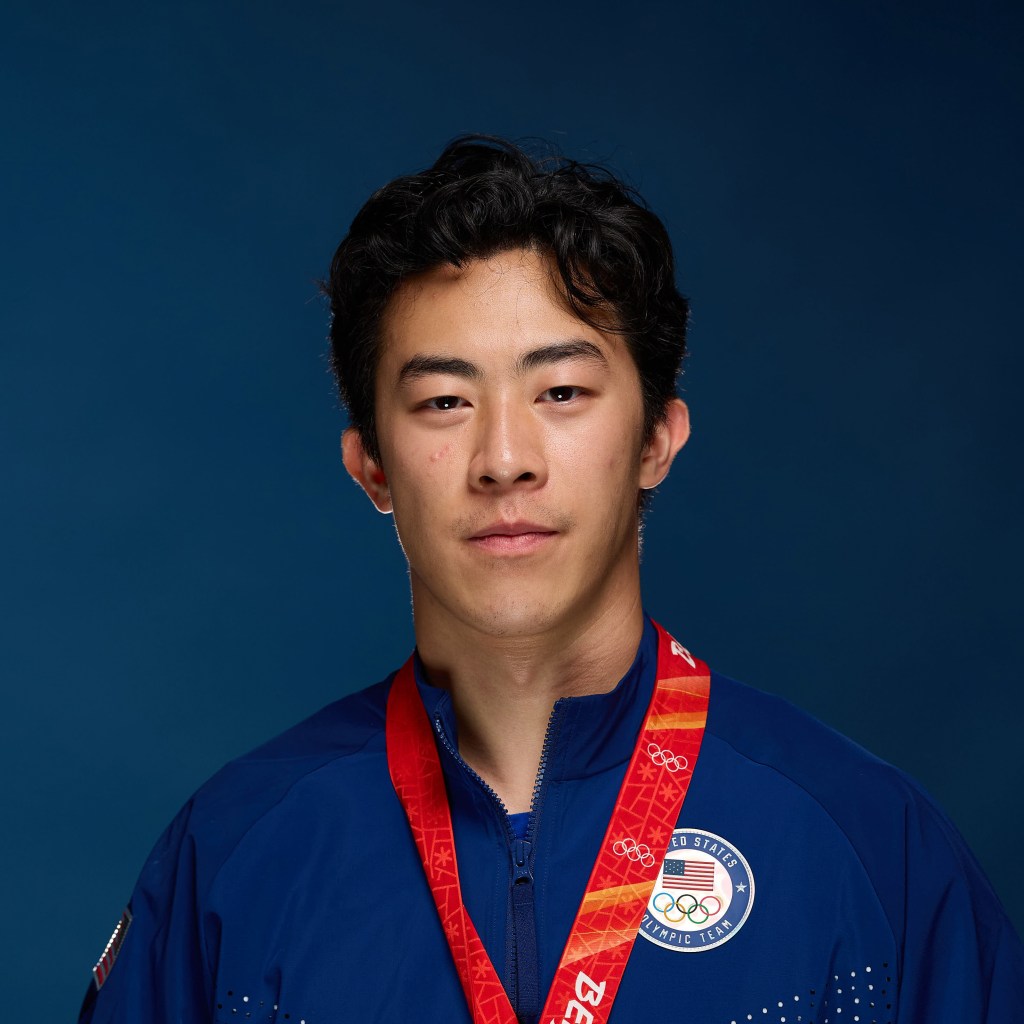 A person wearing a blue jacket with the United States Olympic Team logo, featuring a red medal ribbon  against a solid blue background.
