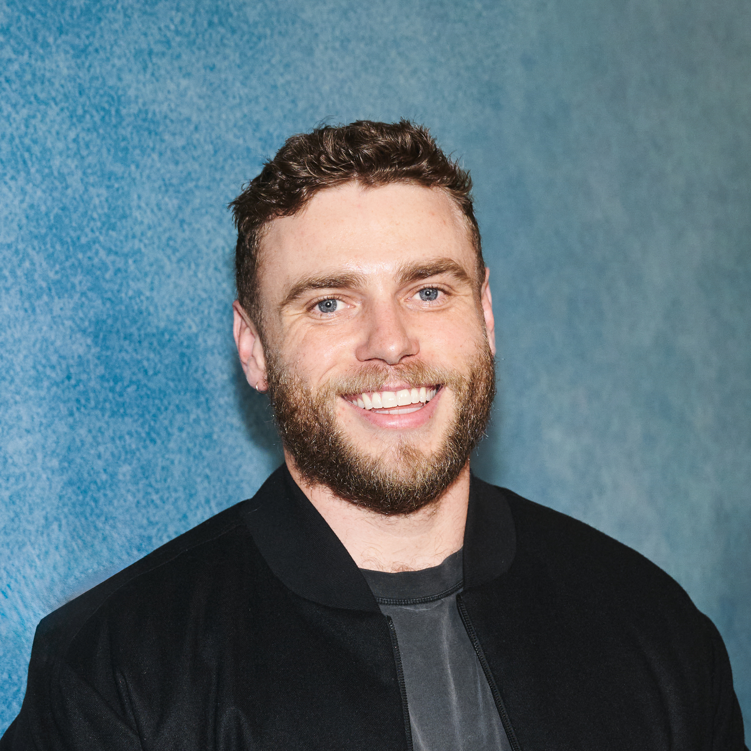 A person wearing a black jacket and gray shirt stands against a textured blue wall background.