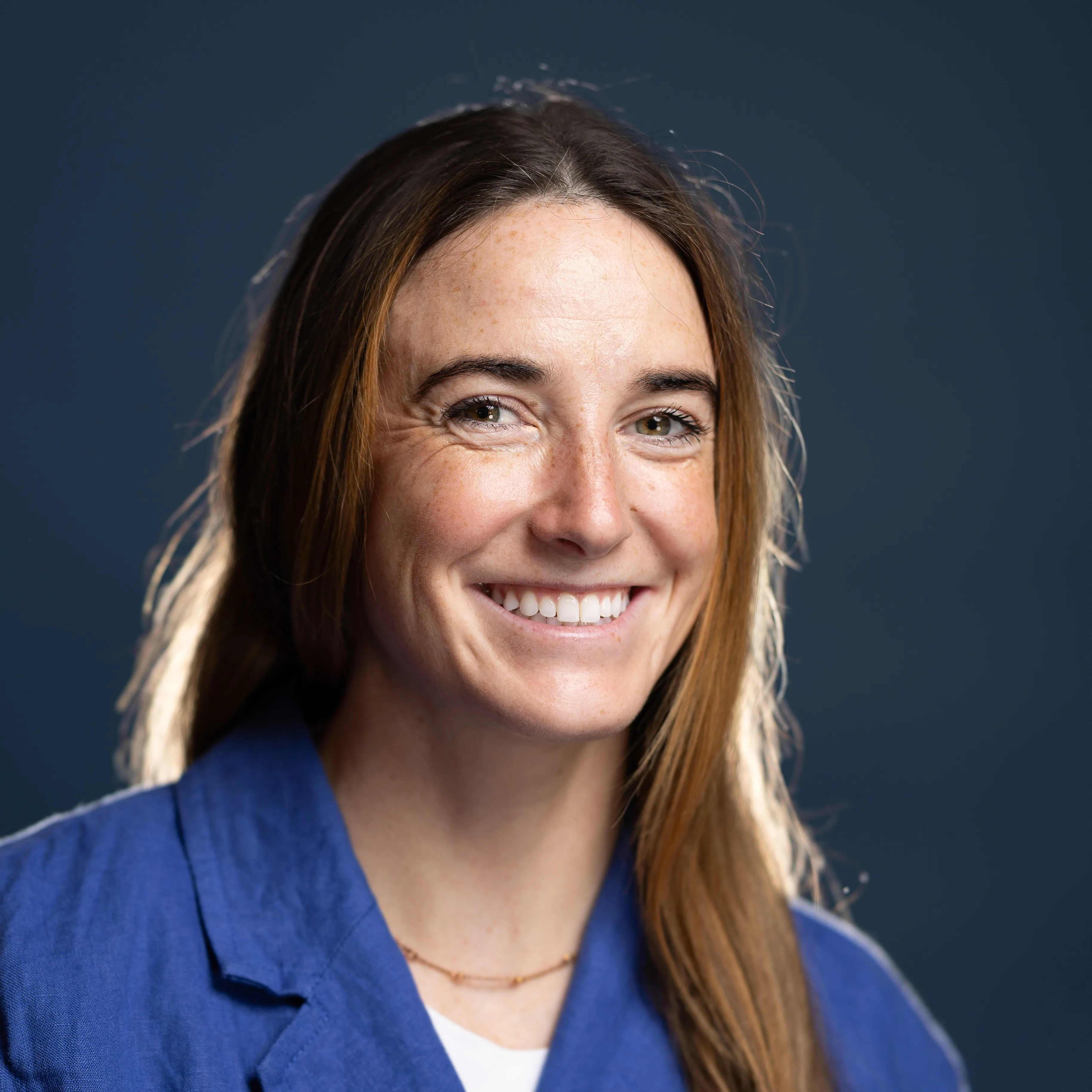 A person with long brown hair wearing a blue collared shirt and a delicate gold necklace, set against a dark blue background.