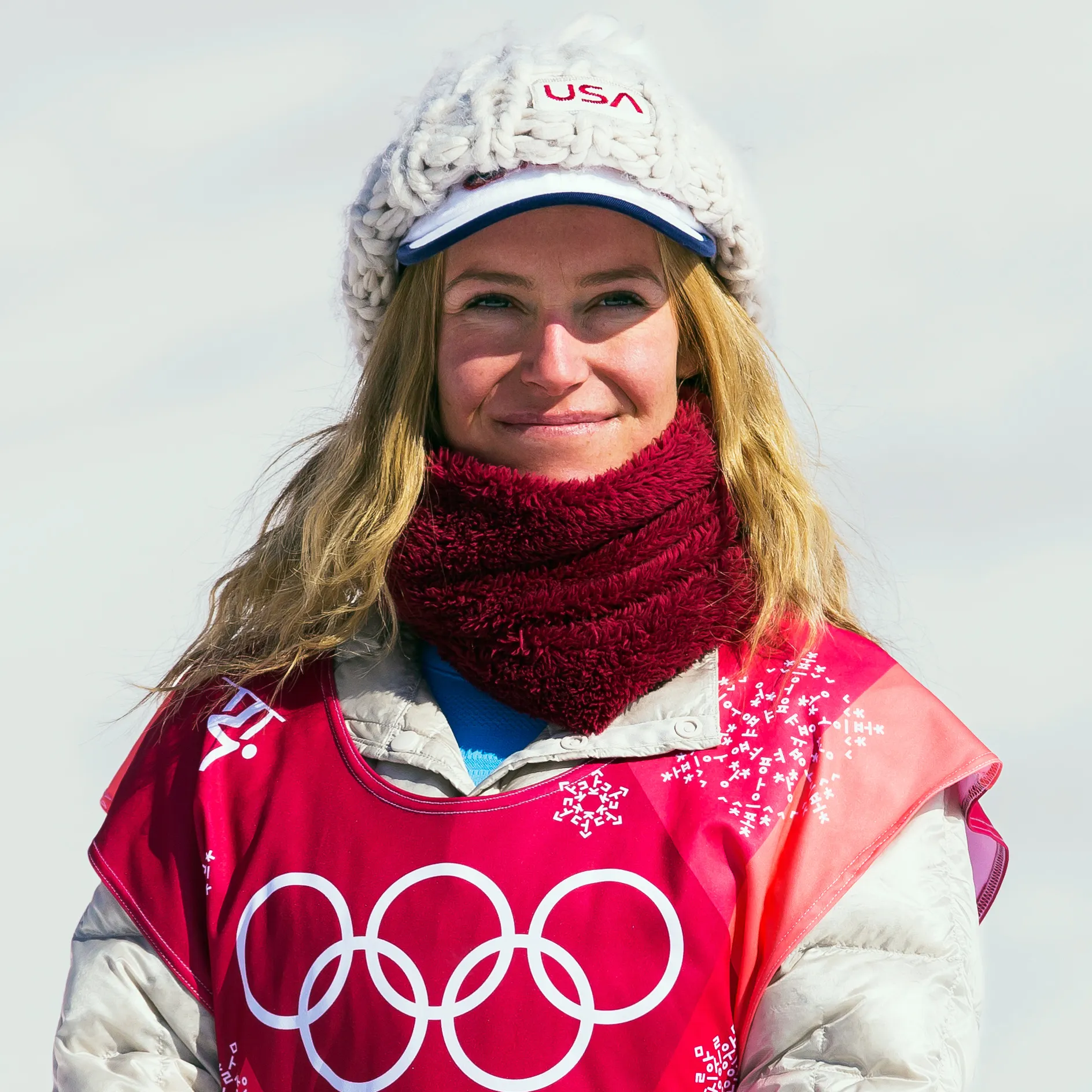 A person wearing a white knit hat with 'USA' written on it, a maroon scarf, and a pink Olympic bib showing the iconic five-ring logo.