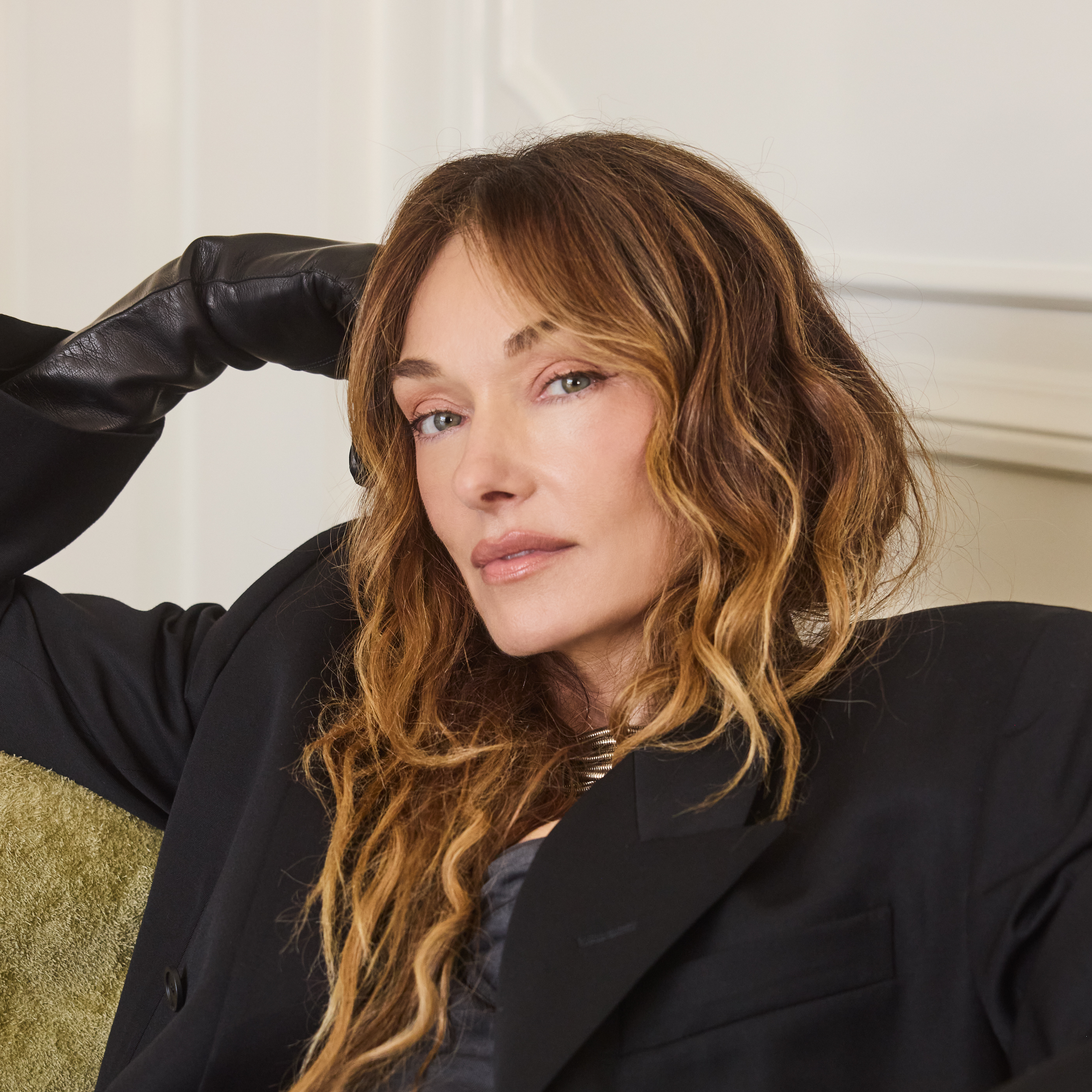 Person with long wavy brown hair wearing a black blazer and leather gloves, reclining on a green upholstered couch in a well-lit room.