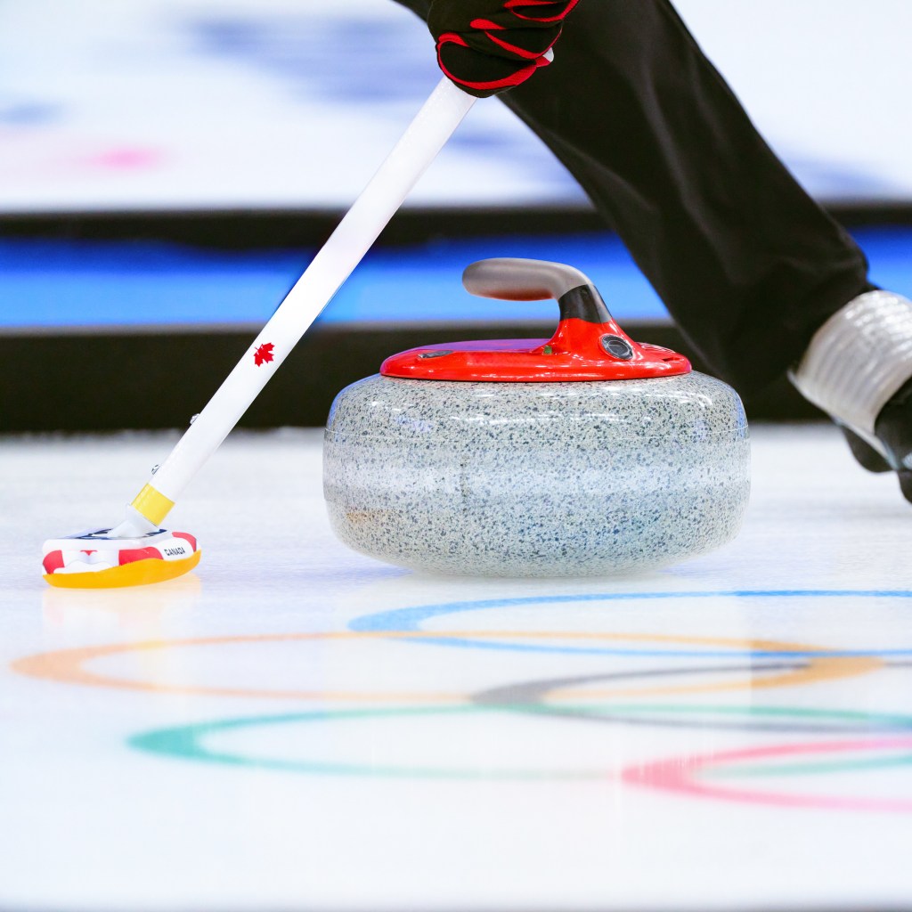Close-up of a curling stone with a red handle sliding on ice, alongside a broom with a Canadian flag emblem during a curling match.