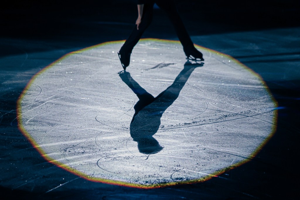 A figure skater gliding gracefully on an ice rink under a spotlight, casting a sharp shadow on the illuminated surface.