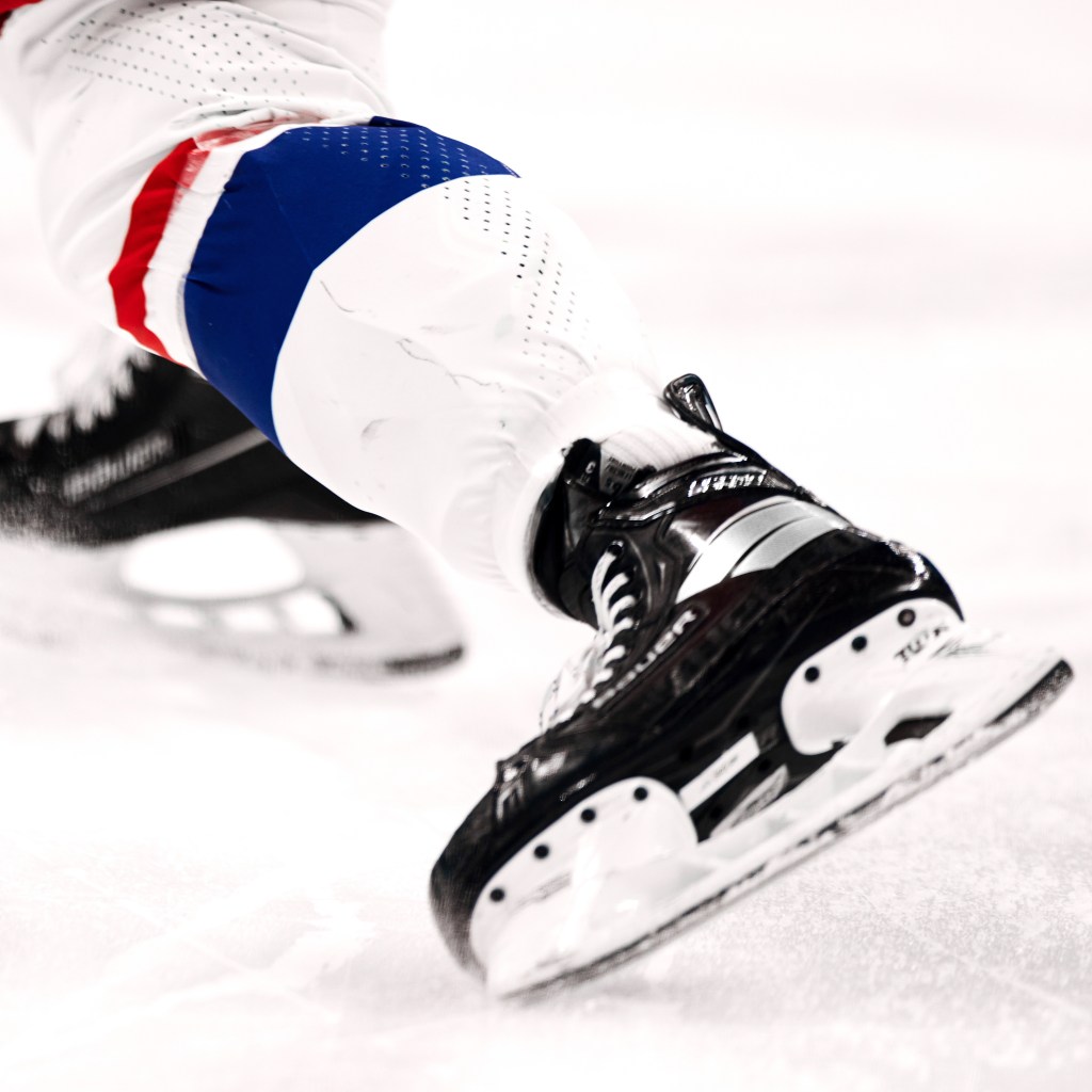 A focused view of a hockey player’s black and white skates cutting through the ice, with blue and red accents on their gear.