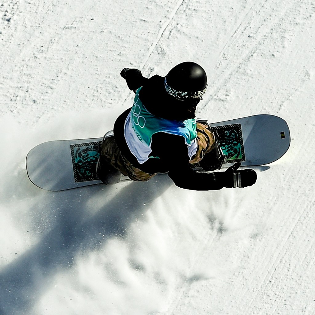 A snowboarder wearing a helmet, goggles, and a colorful jacket glides down a snowy slope, kicking up powder in motion.