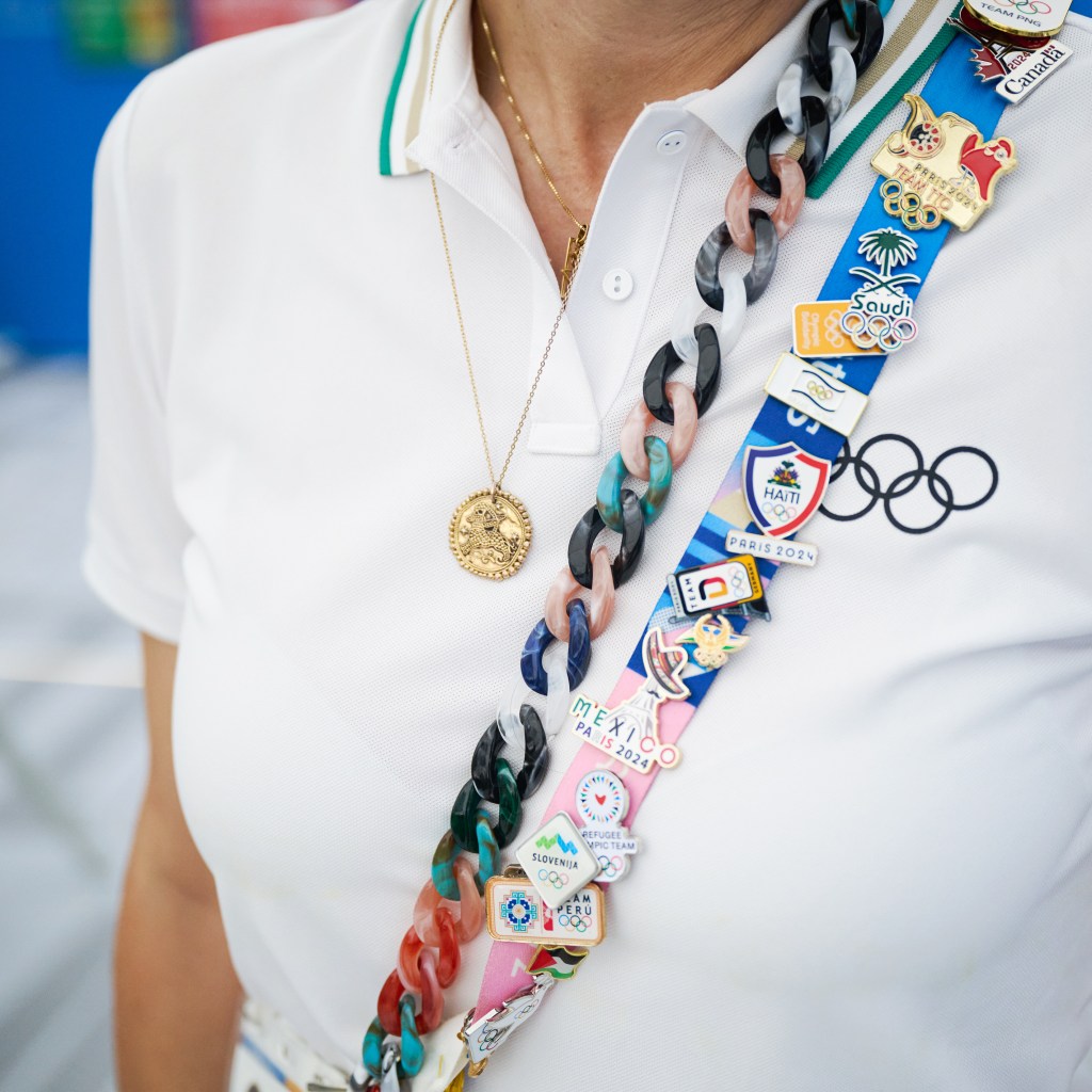 Close-up of a person wearing a white polo shirt with a colorful chain adorned with pins from various countries, including Mexico, Haiti, Slovenia, and Olympic Paris 2024 logos.