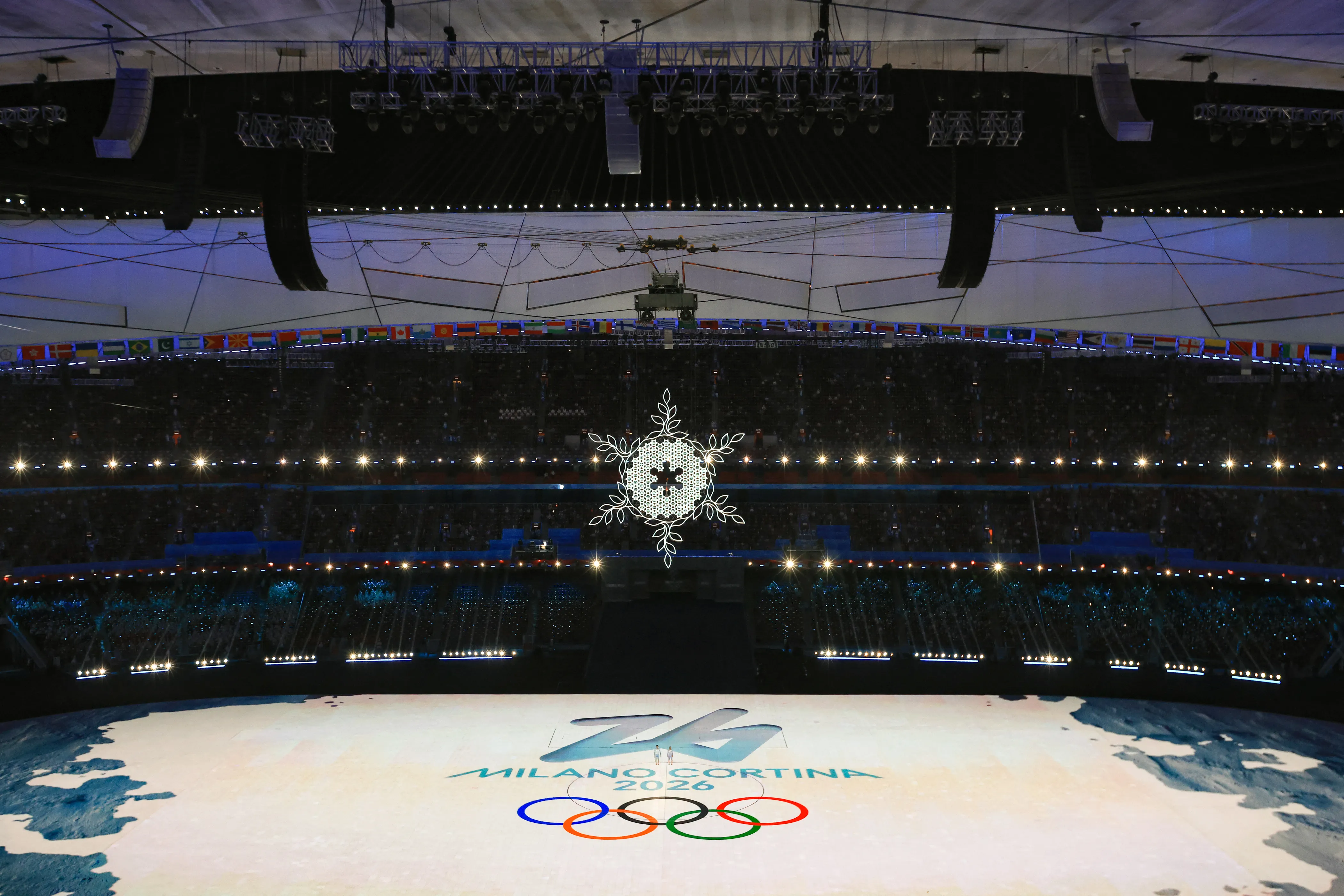 A large indoor stadium illuminated with bright lights, featuring the Milano Cortina 2026 Winter Olympics logo and the Olympic rings projected onto the arena floor. A snowflake chandelier hangs prominently from the ceiling.
