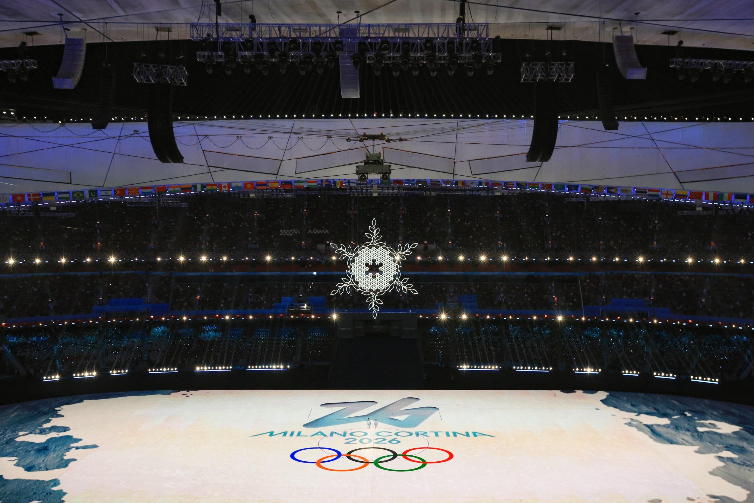 A large indoor stadium illuminated with bright lights, featuring the Milano Cortina 2026 Winter Olympics logo and the Olympic rings projected onto the arena floor. A snowflake chandelier hangs prominently from the ceiling.