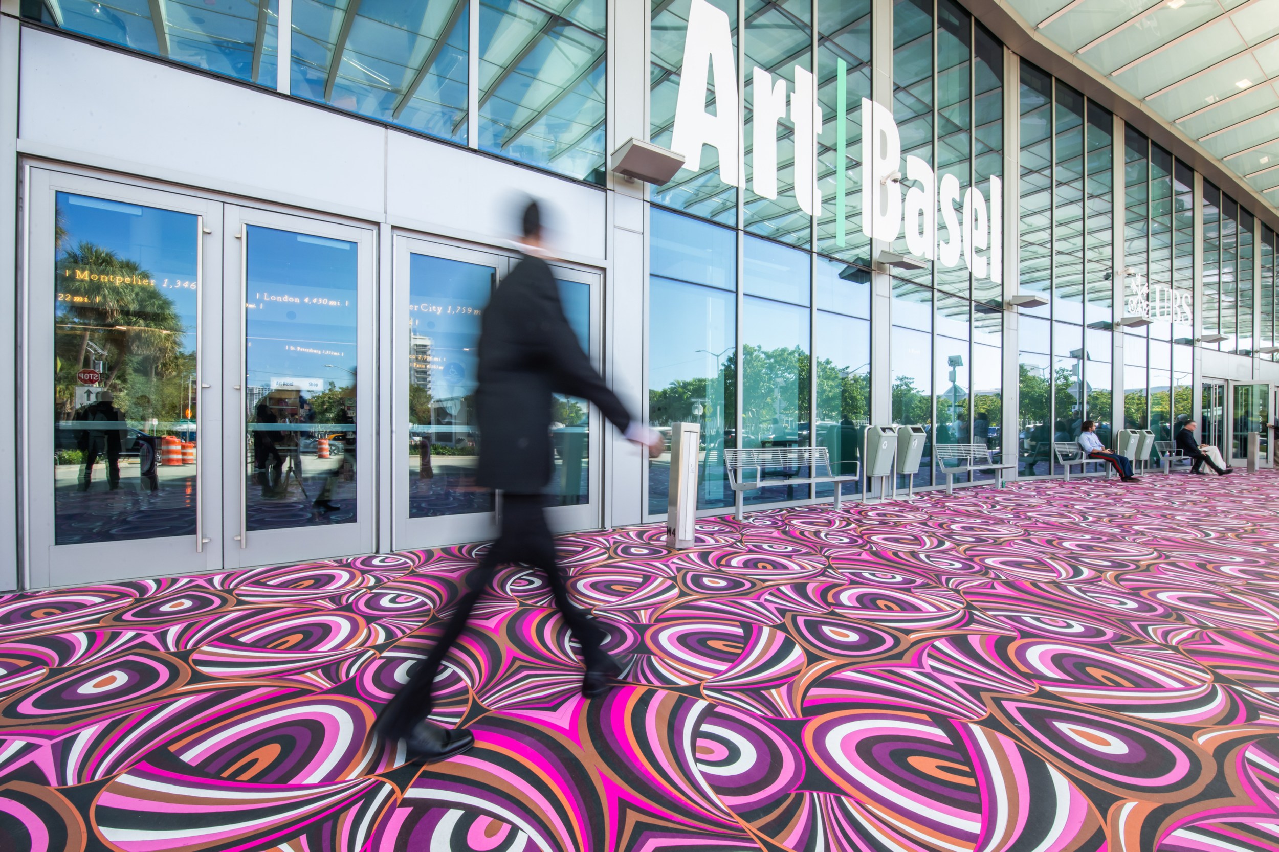 A person walking past the entrance to Art Basel, with colorful pink, purple, and white swirls on the ground and glass doors reflecting greenery in the background.
