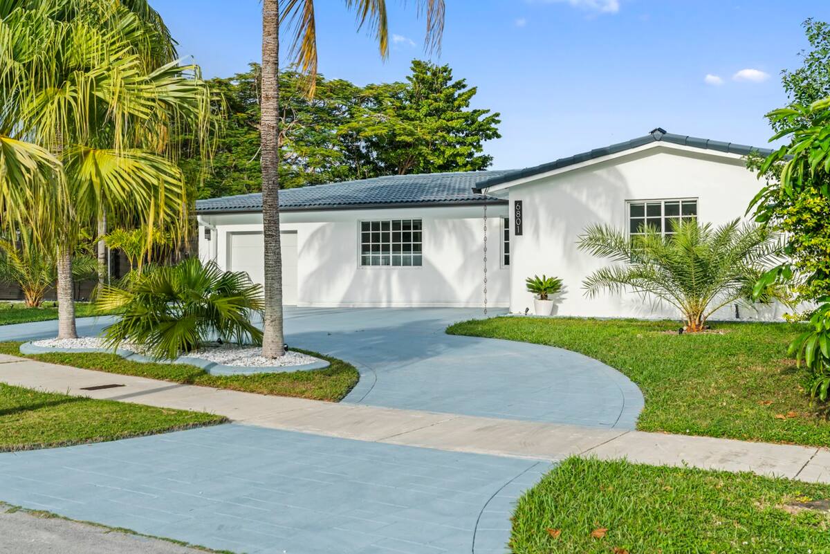 A modern single-story white house with a curved blue driveway, surrounded by palm trees and vibrant green grass on a sunny day.