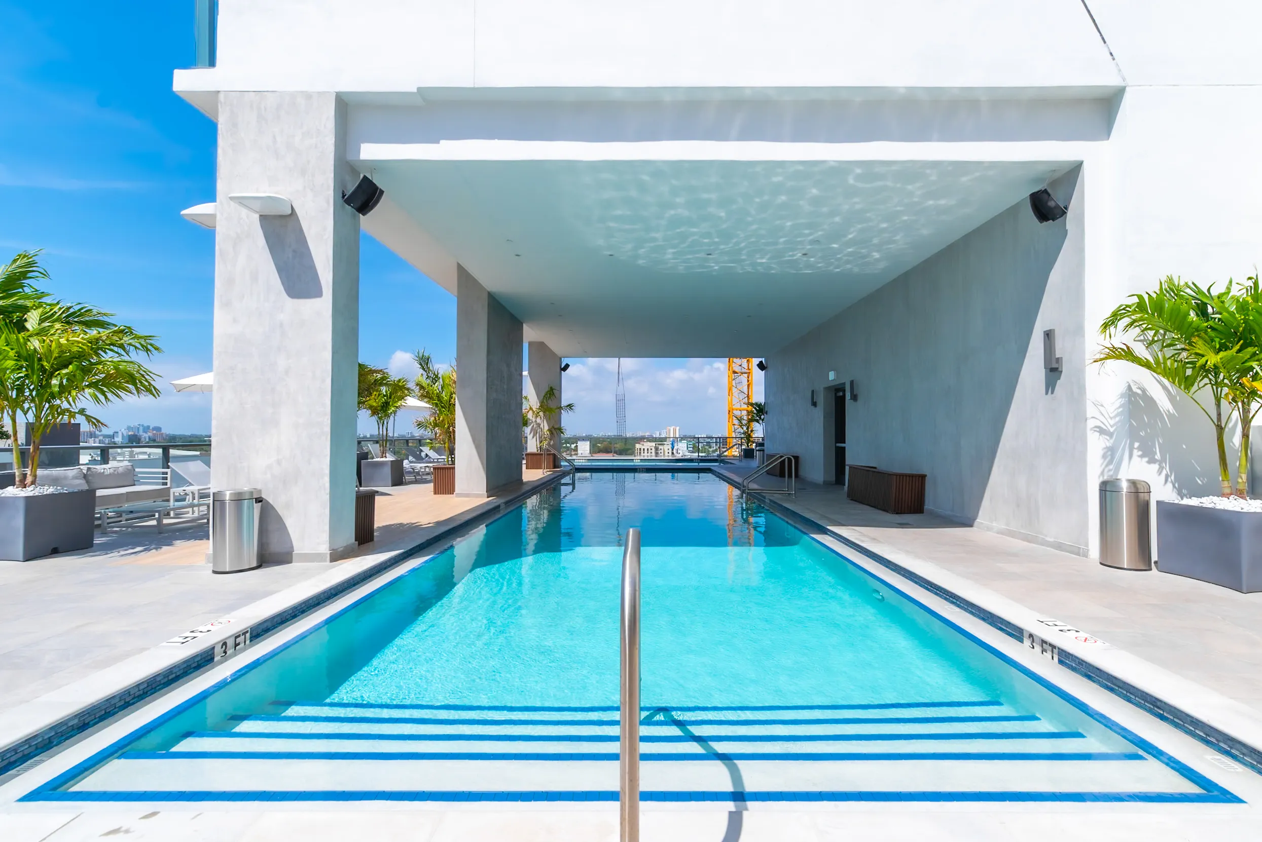 A rooftop swimming pool with clear blue water, surrounded by modern architecture and tropical plants under a bright sky.