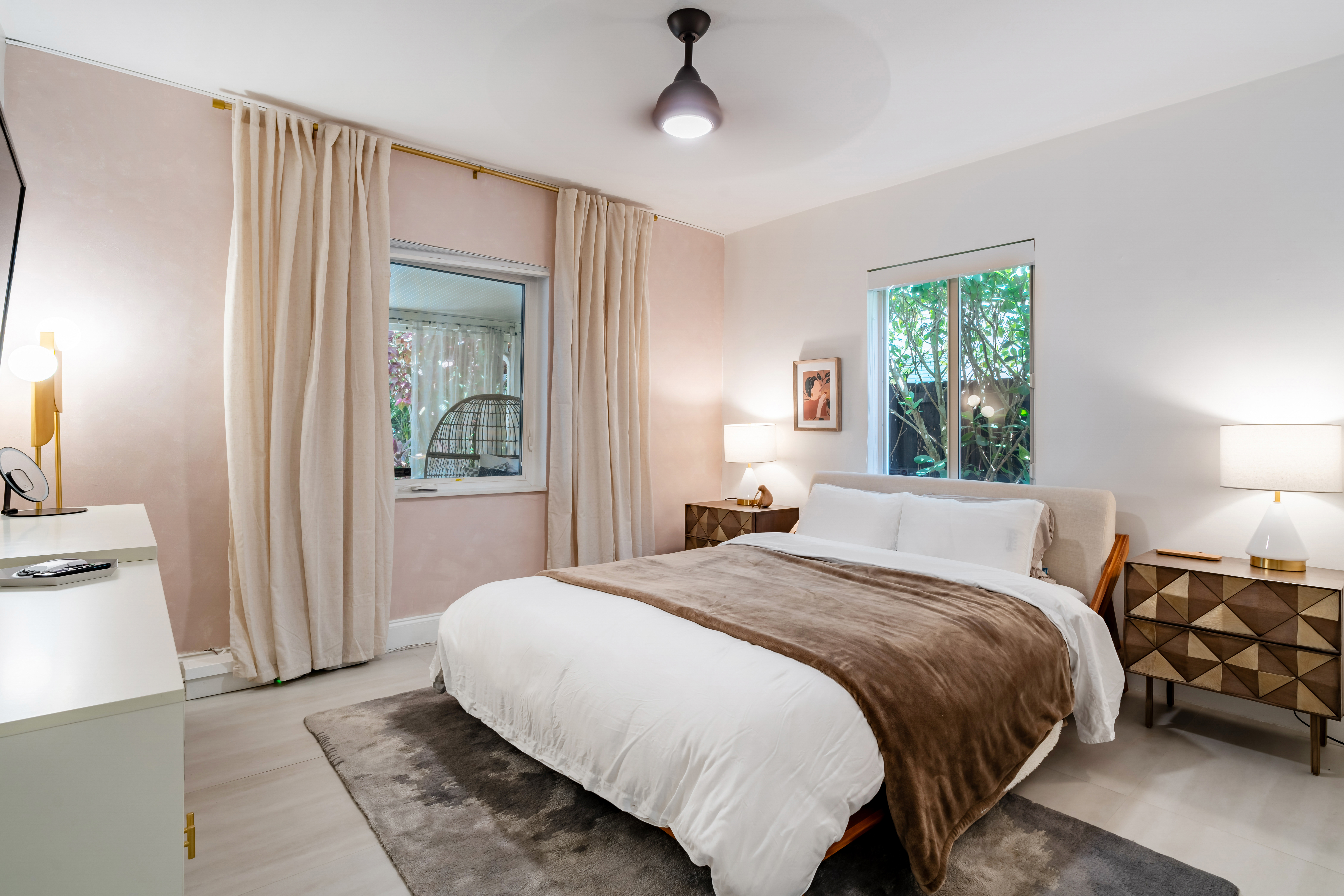Bright and cozy bedroom with a large bed covered in white linens and a brown throw blanket, surrounded by modern furniture and soft lighting, featuring windows with views of greenery outside.