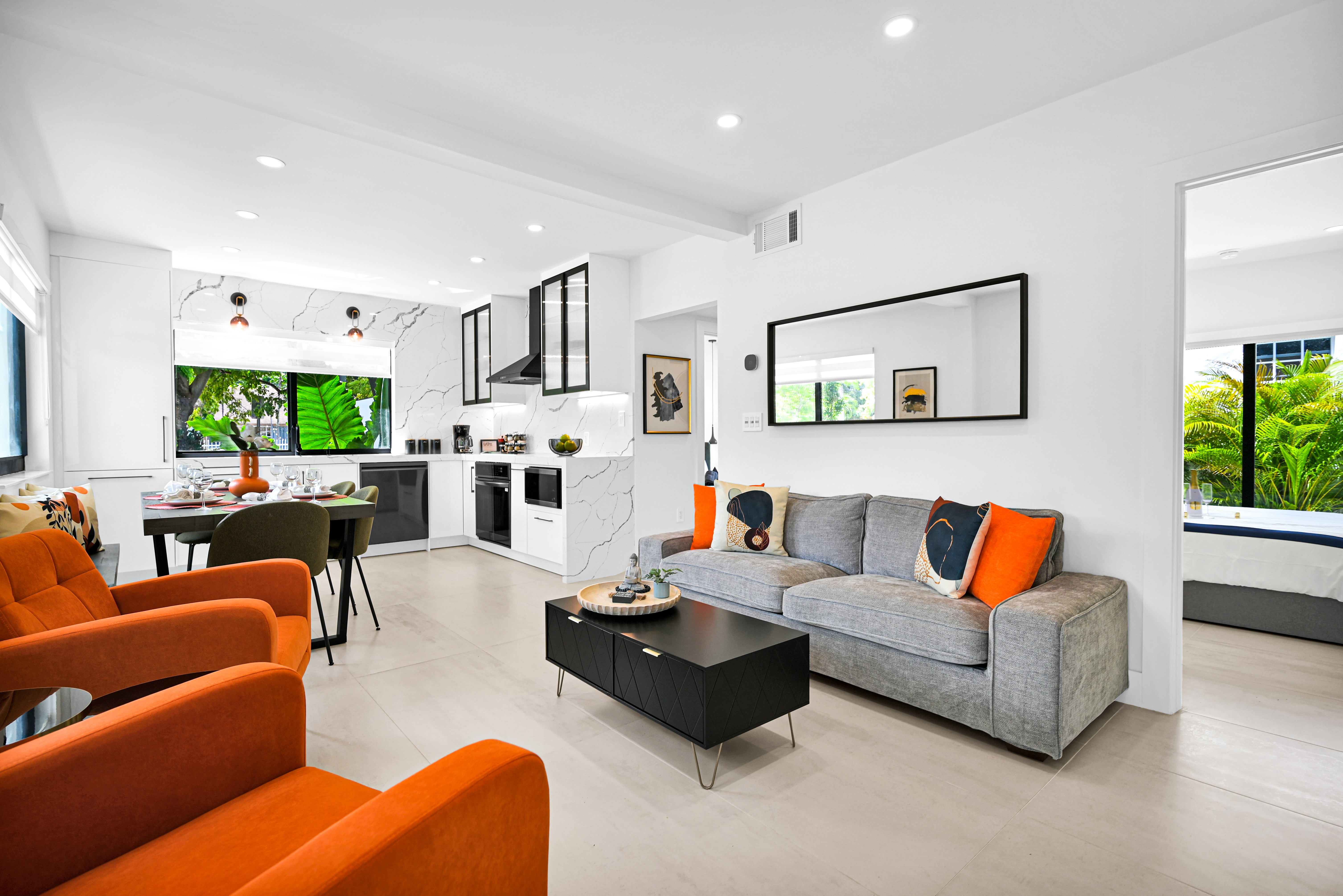A bright and modern living space featuring a gray sofa with orange accent pillows, a black coffee table, a dining table with chairs, and a sleek kitchen in the background. Large windows showcase lush green foliage outside.