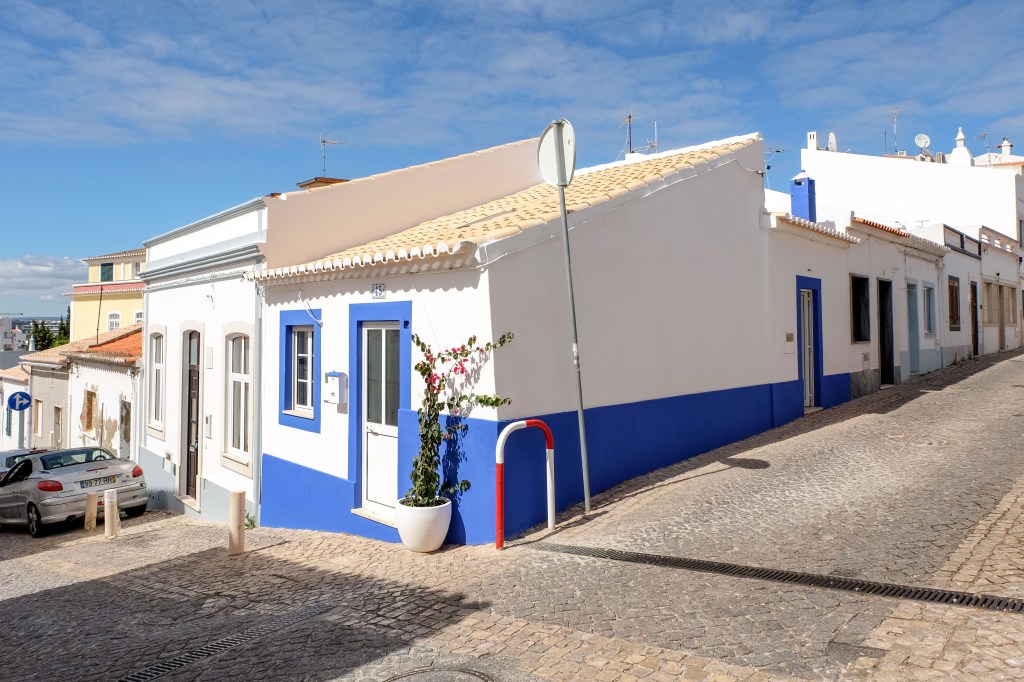 Charming corner house with white and blue walls, terracotta roof tiles, and cobblestone street under a sunny sky.