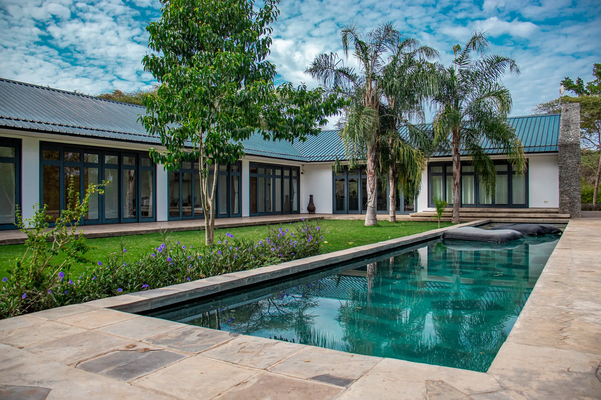 A sleek contemporary villa with large glass doors, lush green landscaping, and a rectangular swimming pool under a partly cloudy sky.