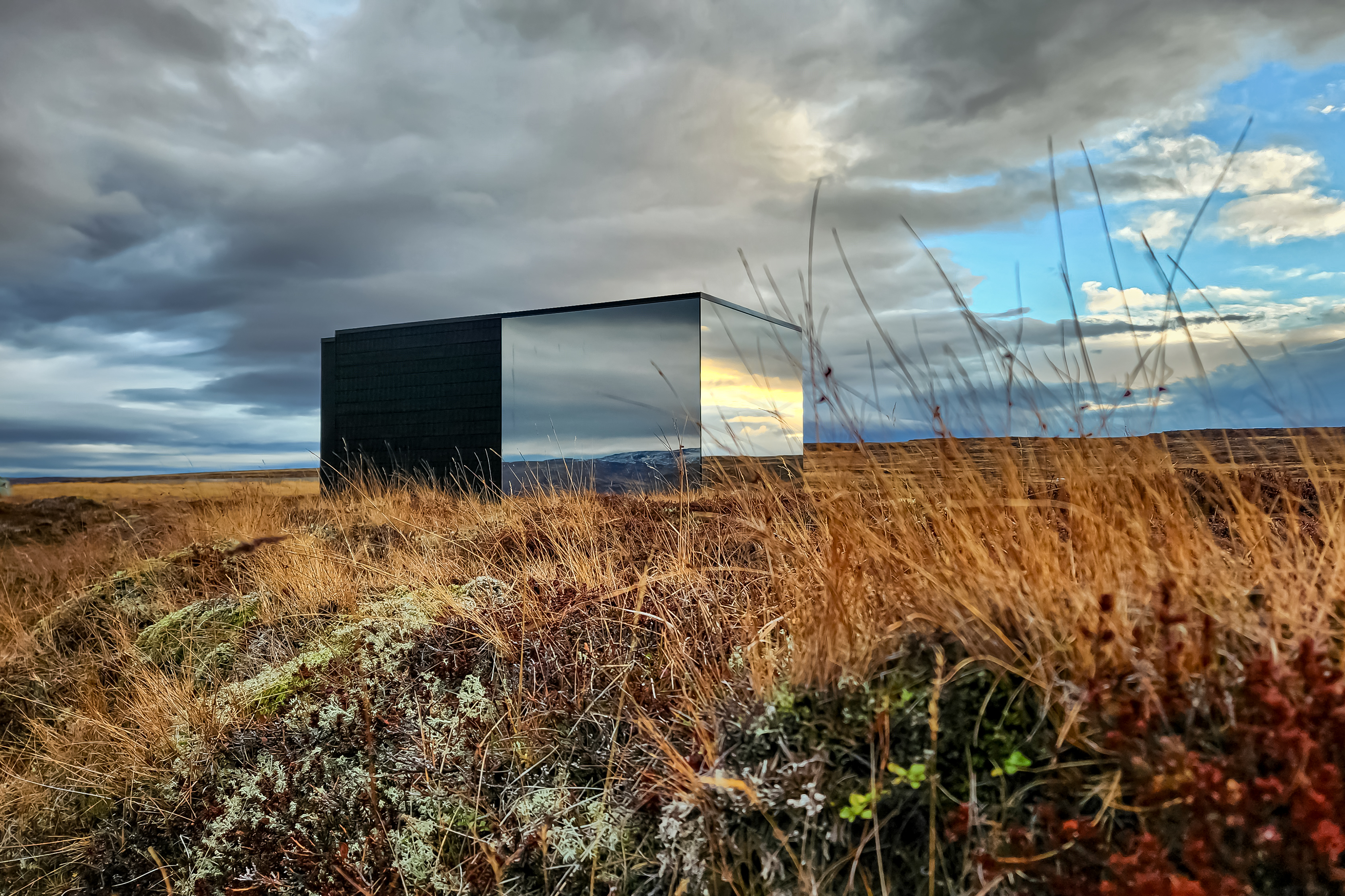 A striking mirrored cube house stands solitary in golden windswept grasslands, reflecting dramatic skies and distant hills.