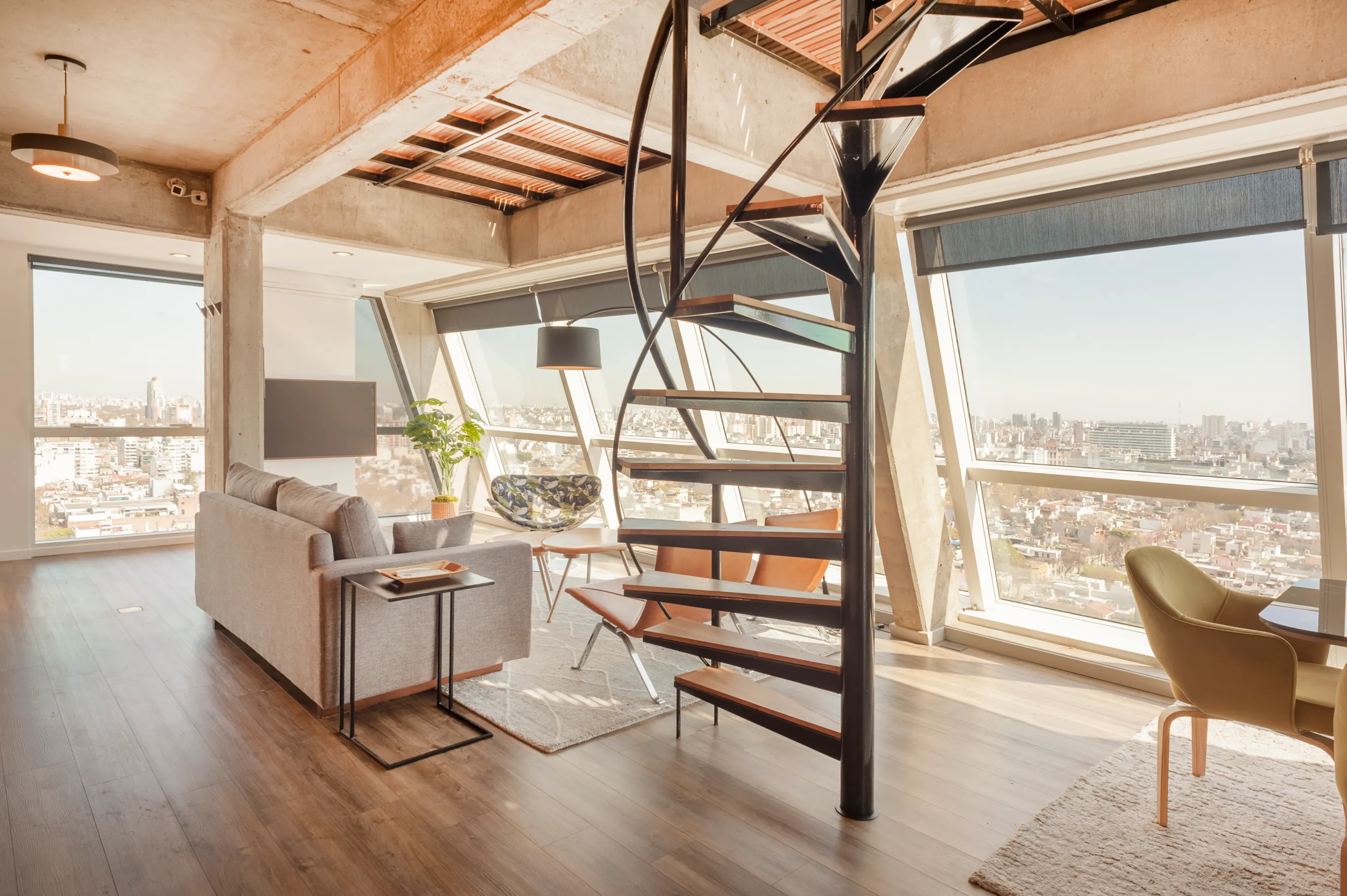 This bright, modern loft features floor-to-ceiling windows with sweeping city views, a stylish spiral staircase, and chic contemporary furnishings in an open-plan living space.