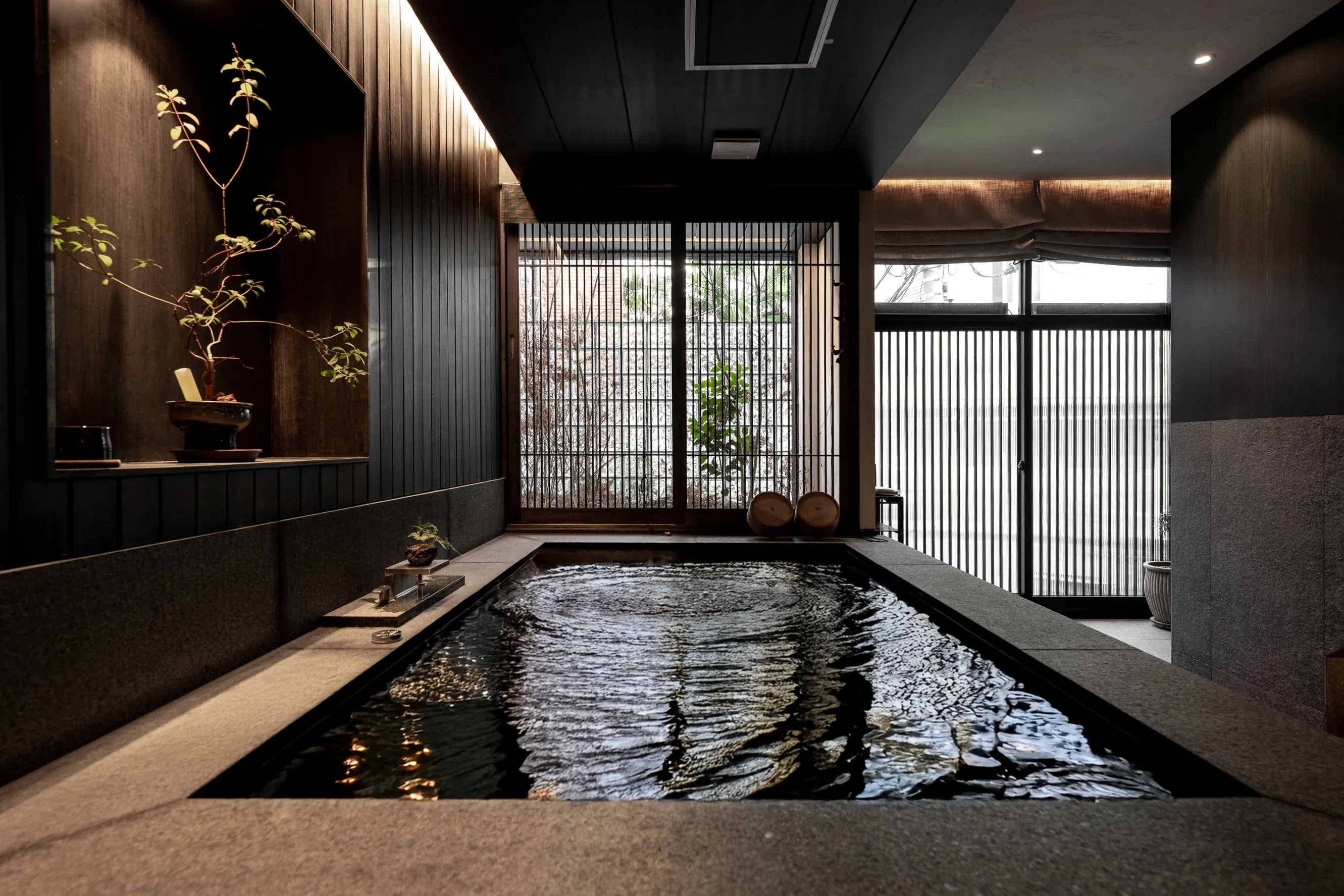A tranquil indoor Japanese-style bath features rippling water, minimalistic decor, and soft natural light streaming through wooden lattice windows