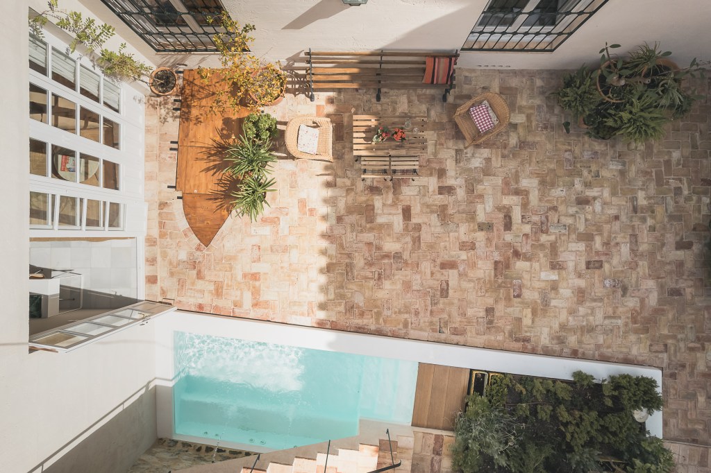 Sunlit Mediterranean-style patio viewed from above, with herringbone tiles, garden plants, comfortable seating, and a small pool.