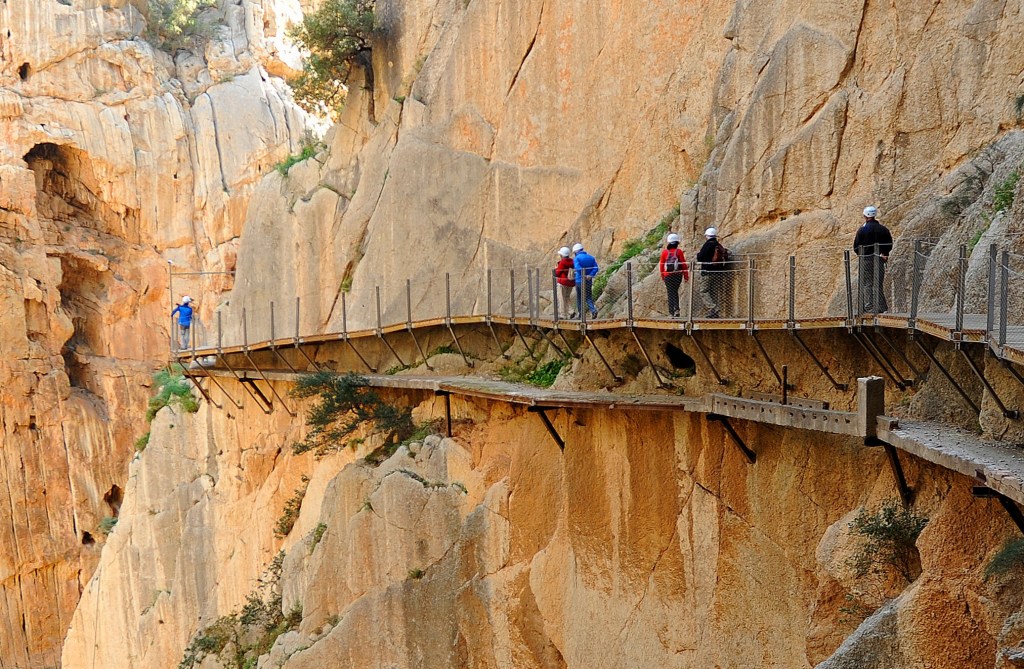 Adventurers walk along a narrow pathway suspended on the side of a rocky canyon.