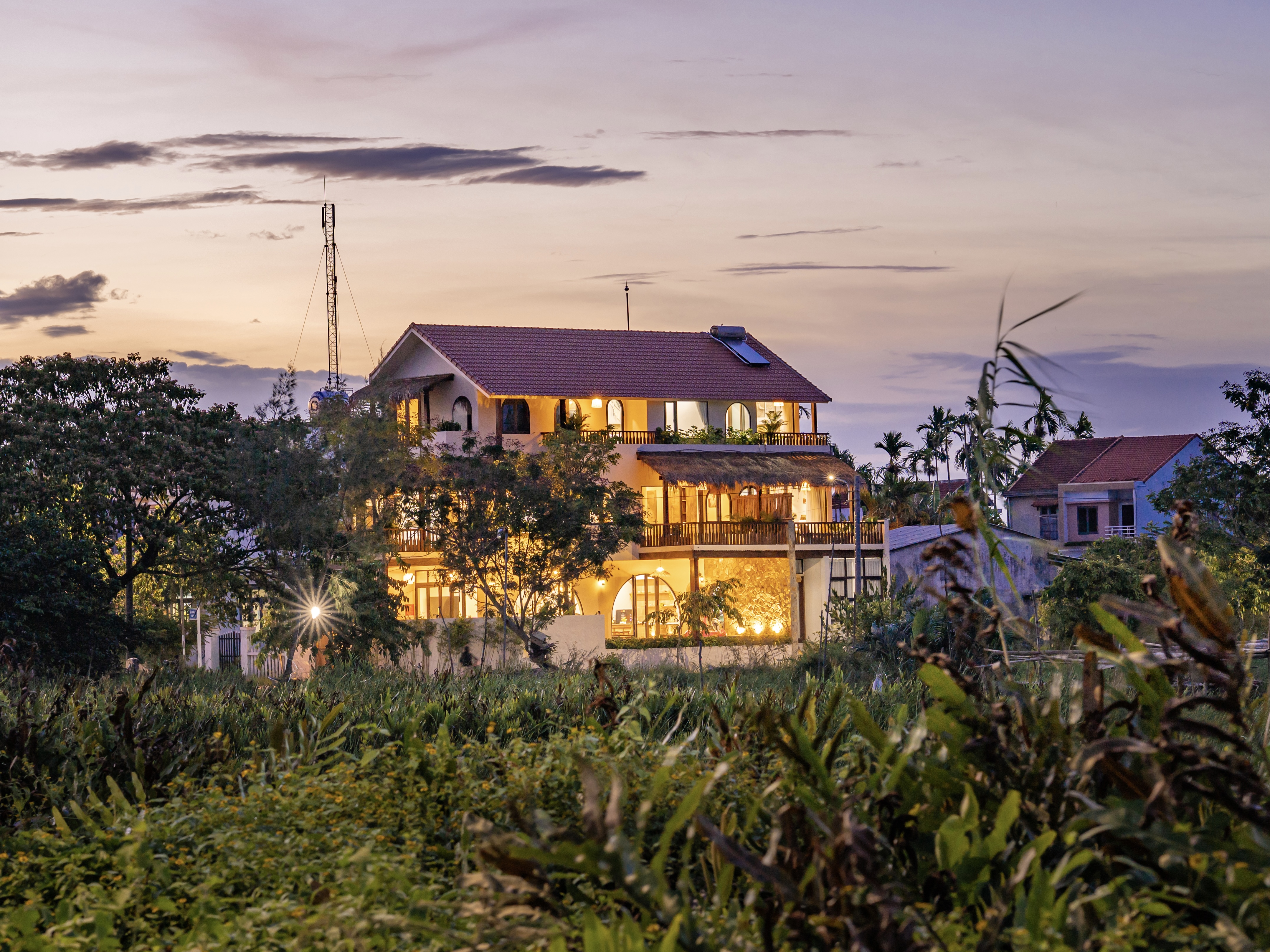 Spacious Mediterranean-style villa glowing warmly at dusk, surrounded by lush greenery and open sky.