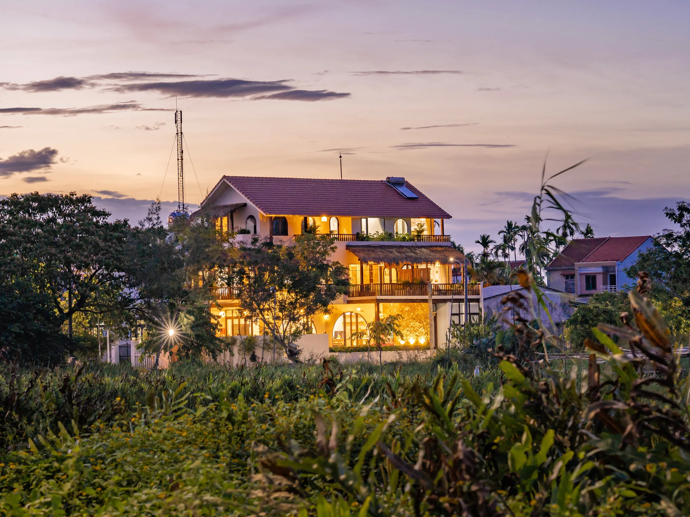 Spacious Mediterranean-style villa glowing warmly at dusk, surrounded by lush greenery and open sky.