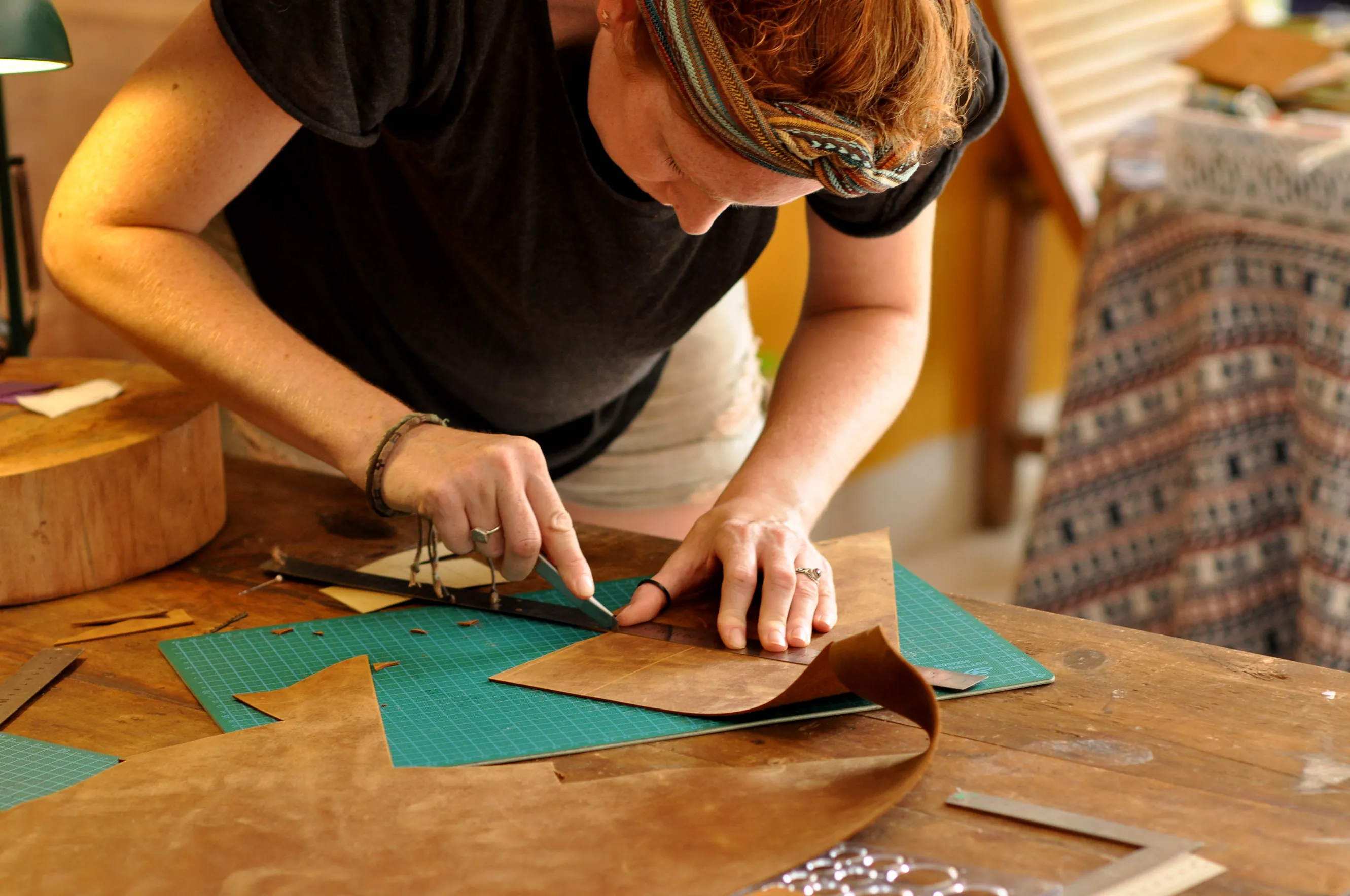 A person is carefully cutting a piece of brown leather on a worktable using a precision knife and ruler, surrounded by crafting tools and materials.