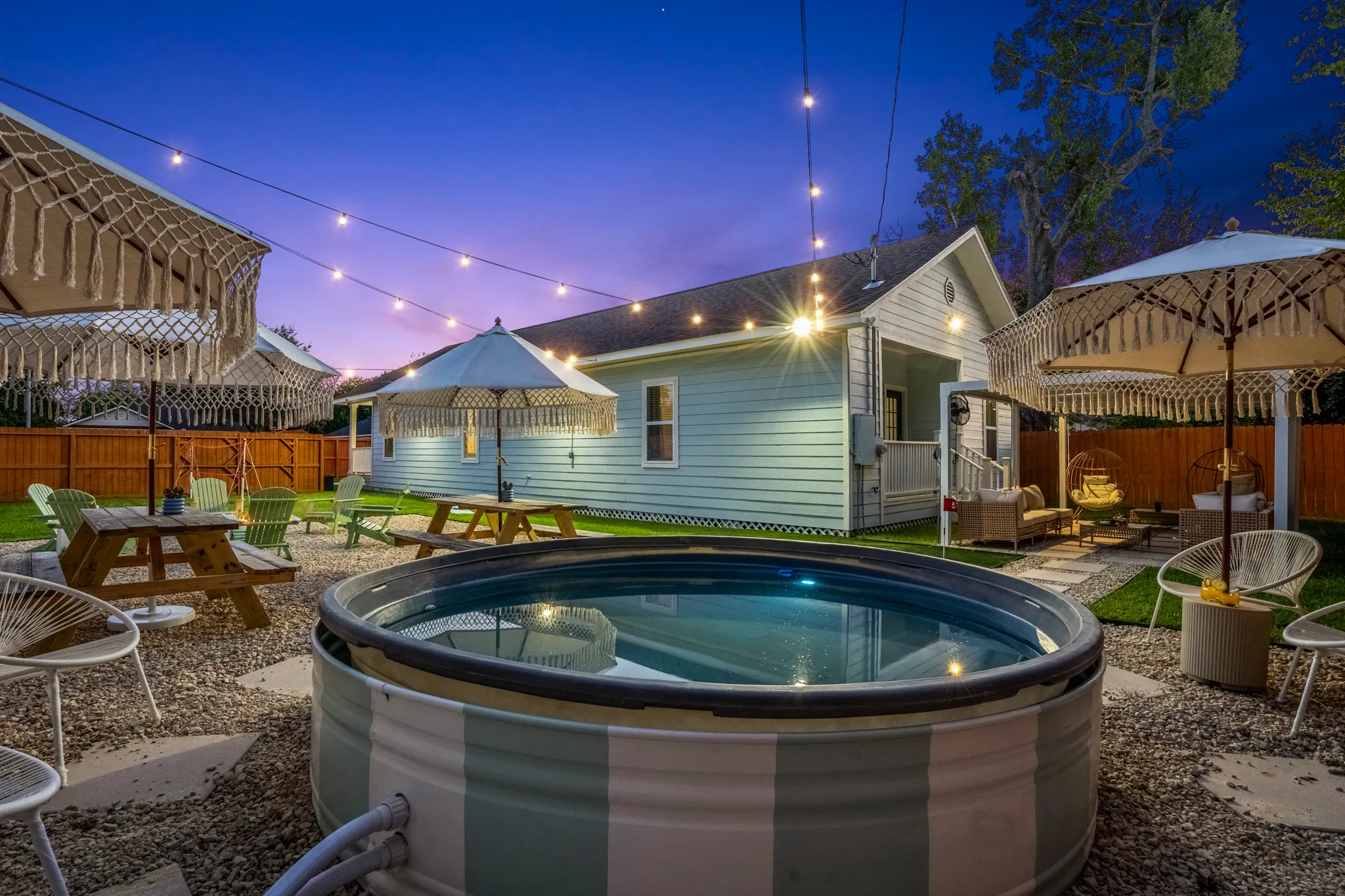 Charming backyard retreat at dusk featuring string lights, picnic tables, macrame umbrellas, and a round soaking pool.