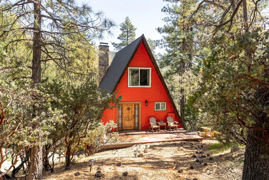 Bright red A-frame cabin with a rustic wooden door, set in a sun-dappled forest clearing.