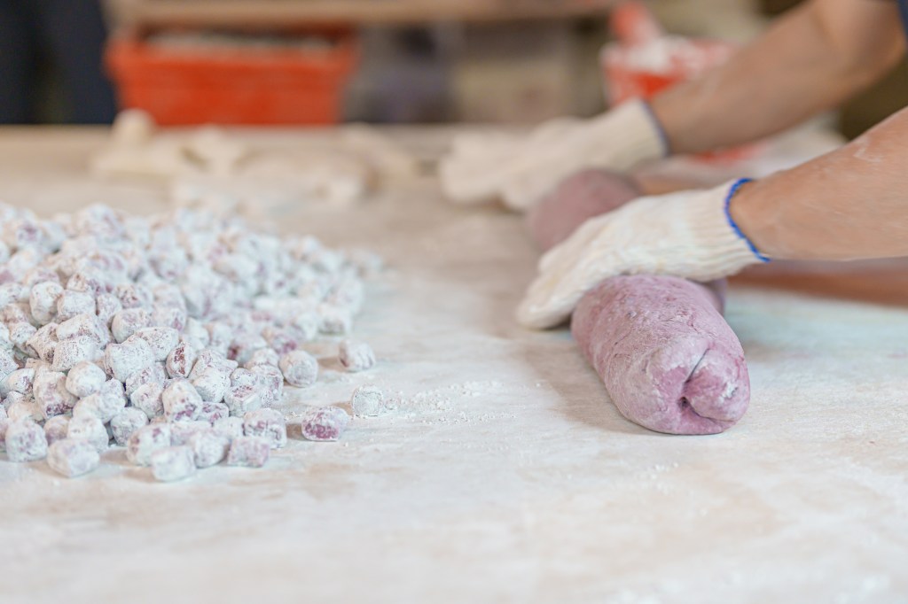 Gloved hands shape a long roll of purple dough on a floured surface next to small, flour-dusted dough pieces.