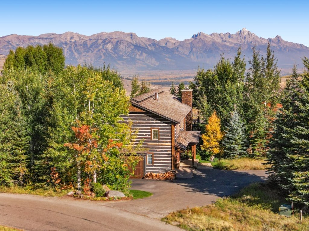 A classic log cabin sits among lush trees with colorful foliage, set against a stunning mountain backdrop under a clear blue sky.