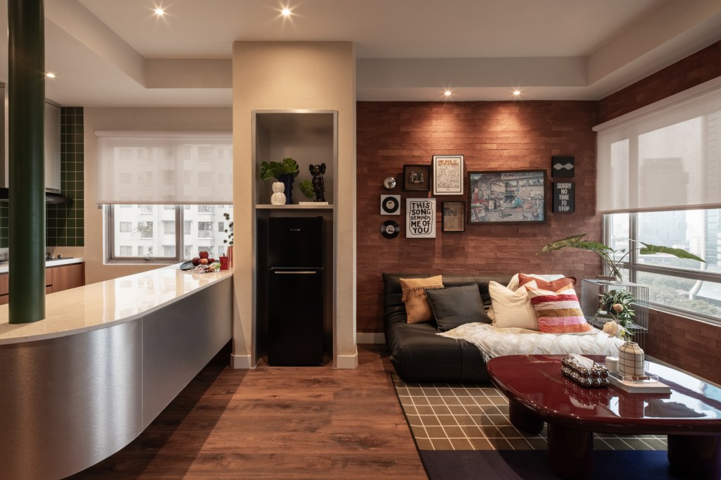 A modern, cozy living space features a sleek kitchen with green tiles, a stylish lounge with framed art and plush pillows, and large windows overlooking city buildings.
