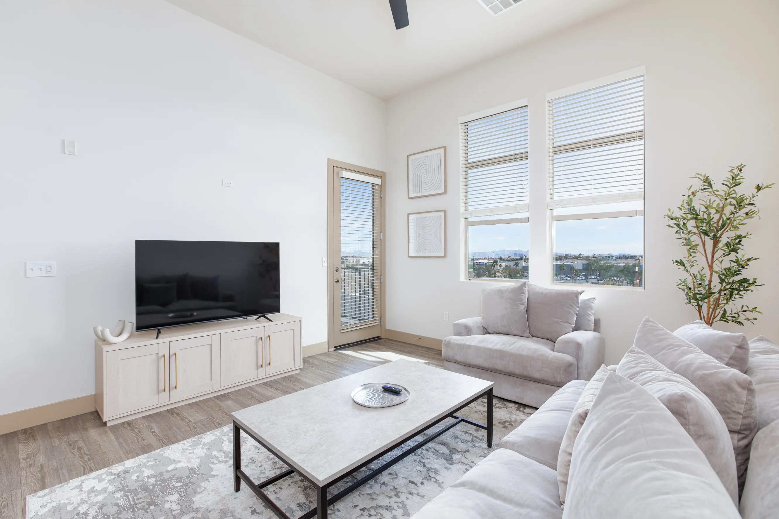 A bright modern living room features plush gray sofas, a large TV on a light wood console, expansive windows with city views, and minimalist decor.
