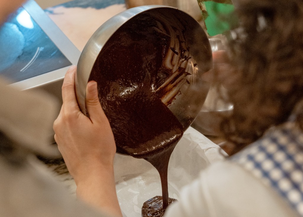 Chocolate batter is being poured from a metal bowl into a baking tin lined with parchment paper.