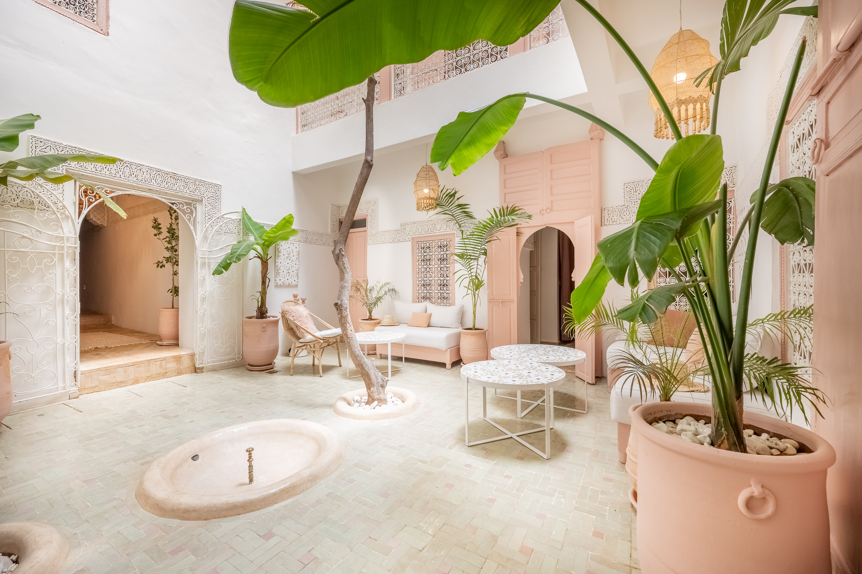 A serene courtyard with pastel pink walls, lush greenery, and ornate archways evokes a peaceful, Moroccan-inspired retreat.