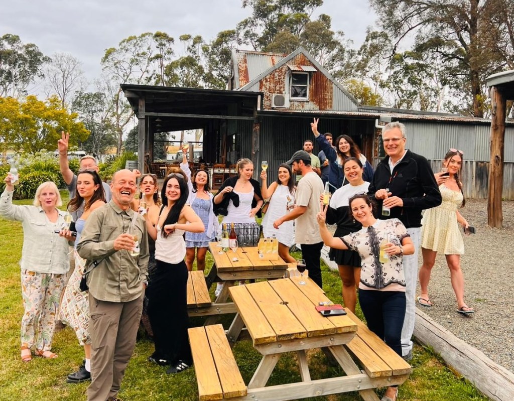A group of people socialize and pose around picnic tables outdoors in front of rustic barn-style buildings.